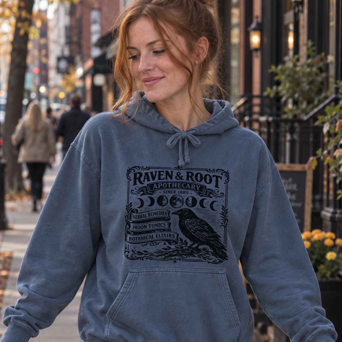 A woman walking on a city sidewalk wearing a faded slate blue hoodie with a black graphic print that reads 'RAVEN & ROOTS' and features a raven illustration. She is also wearing light-wash jeans.