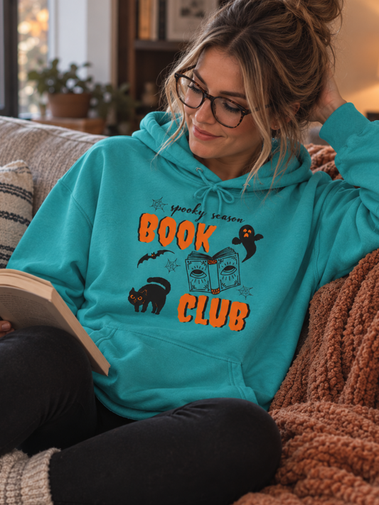 A woman wearing a bright turquoise hoodie with a 'Grim Grinning Book Club' graphic sits on a cozy couch reading a book in a warm, living room setting.