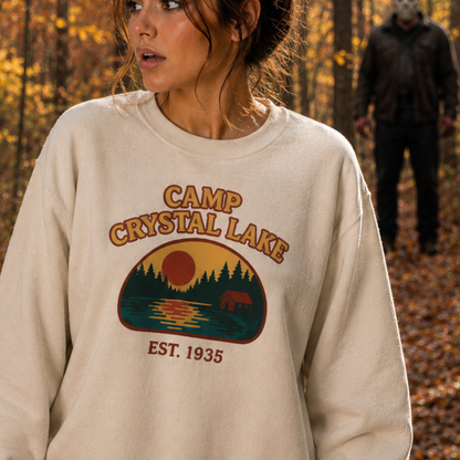 A woman wearing a cream-colored crewneck sweatshirt with a 'Camp Crystal Lake' graphic on the front, set against an autumn forest background with a blurred figure in the distance.