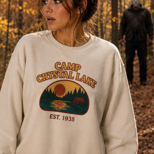 A woman wearing a cream-colored crewneck sweatshirt with a 'Camp Crystal Lake' graphic on the front, set against an autumn forest background with a blurred figure in the distance.