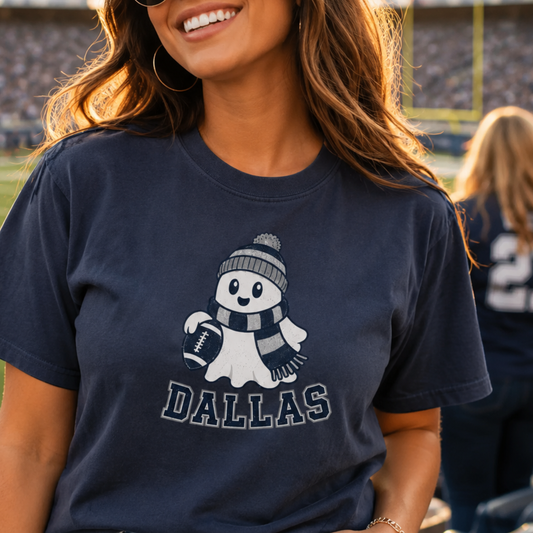 A woman wearing a navy blue graphic t-shirt with a cartoon snowman wearing a beanie and scarf holding a football, with the word 'DALLAS' printed below. The background shows a blurred stadium setting.