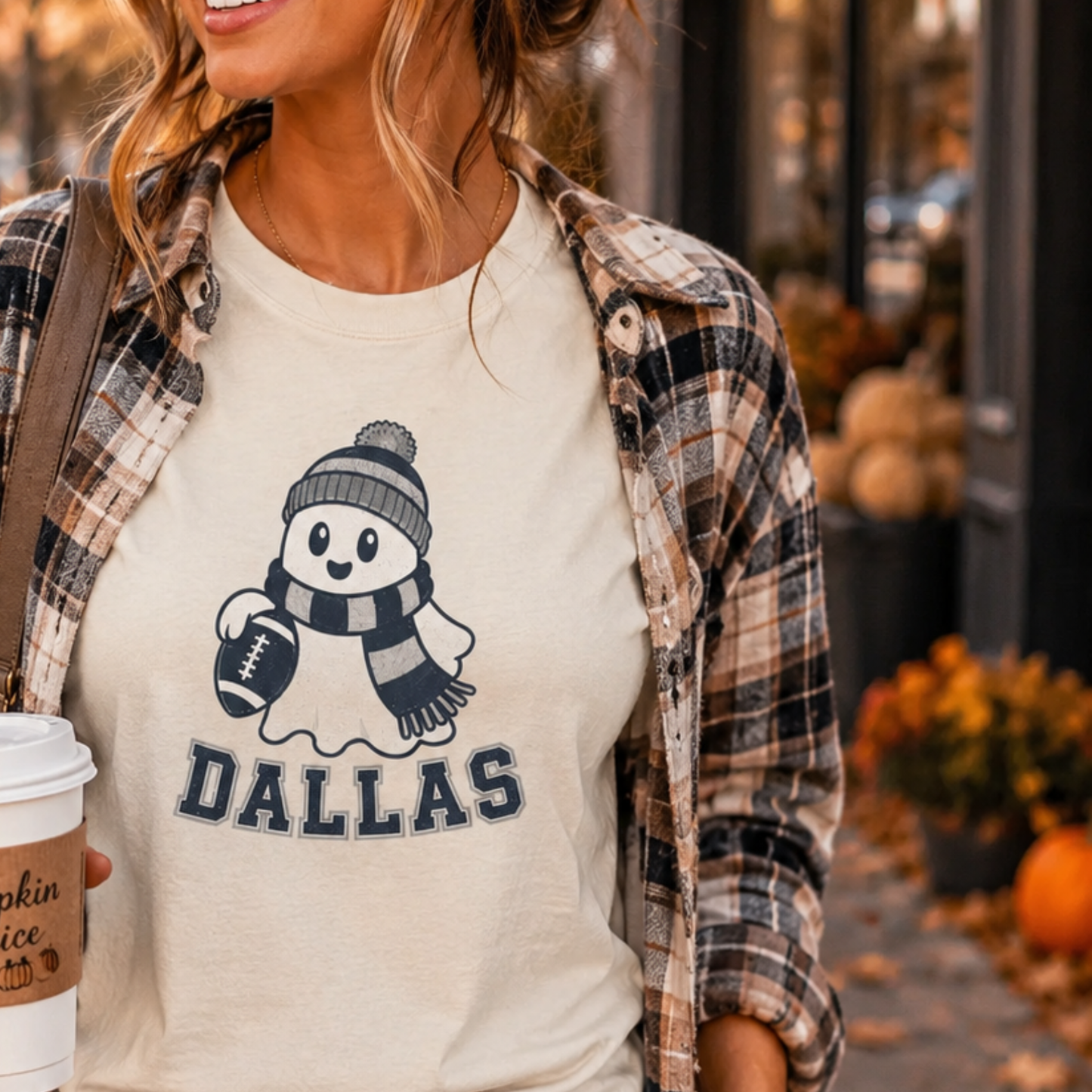A woman wearing a cream graphic t-shirt with a navy blue snowman and the word 'DALLAS' underneath, layered with an open brown and beige plaid flannel shirt. She is holding a coffee cup in a fall-themed outdoor setting with pumpkins.