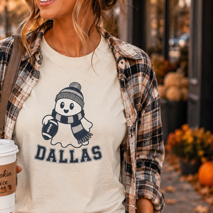 A woman wearing a cream graphic t-shirt with a navy blue snowman and the word 'DALLAS' underneath, layered with an open brown and beige plaid flannel shirt. She is holding a coffee cup in a fall-themed outdoor setting with pumpkins.