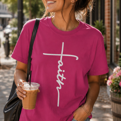 A woman wearing a bright pink t-shirt with a white script graphic spelling 'faith' across the chest, holding an iced coffee on a sunny outdoor street.
