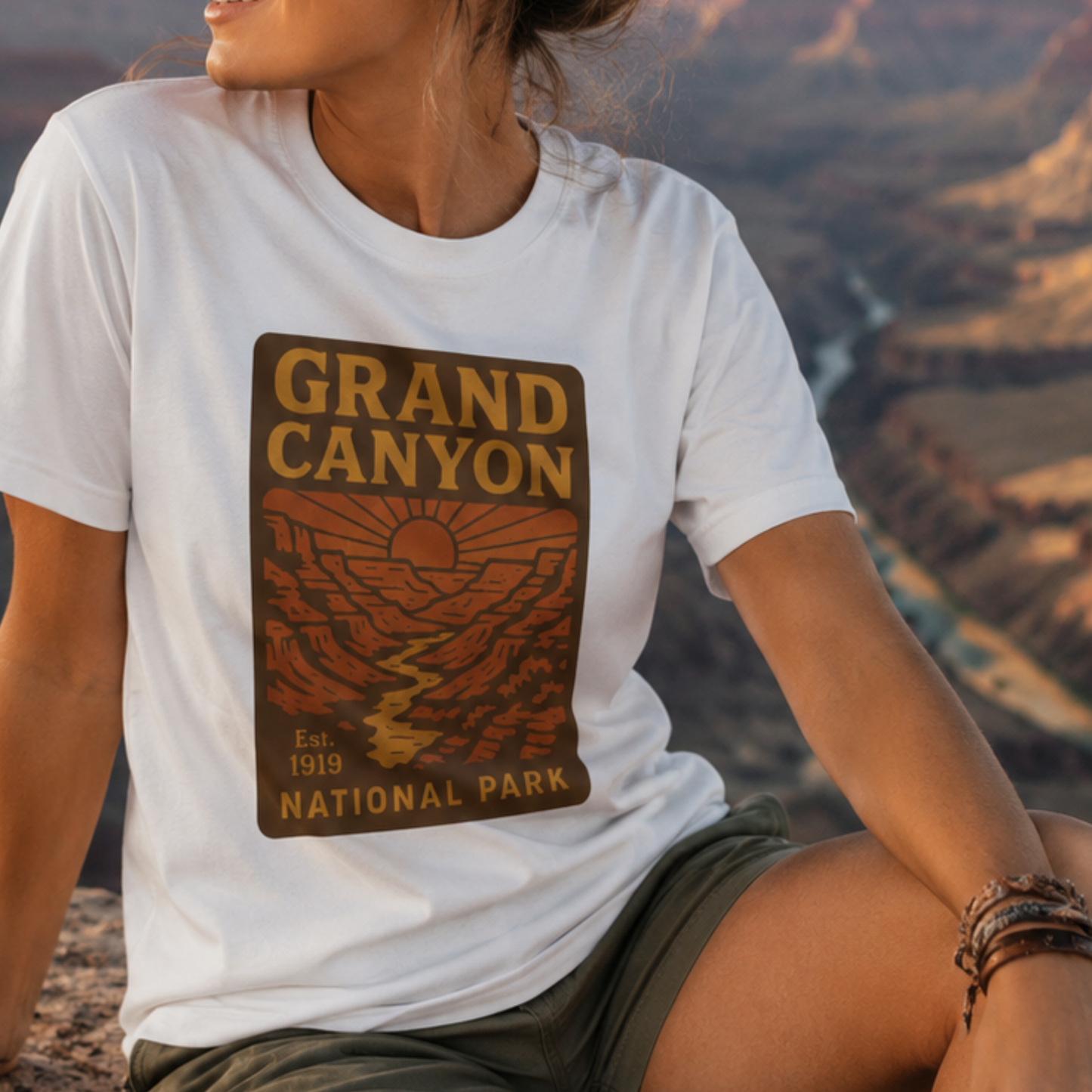 A woman wearing a white t-shirt with a Grand Canyon National Park graphic print, sitting outdoors with a canyon landscape in the background.