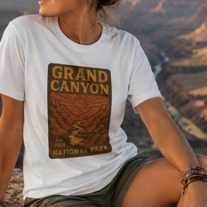 A woman wearing a white t-shirt with a Grand Canyon National Park graphic print, sitting outdoors with a canyon landscape in the background.
