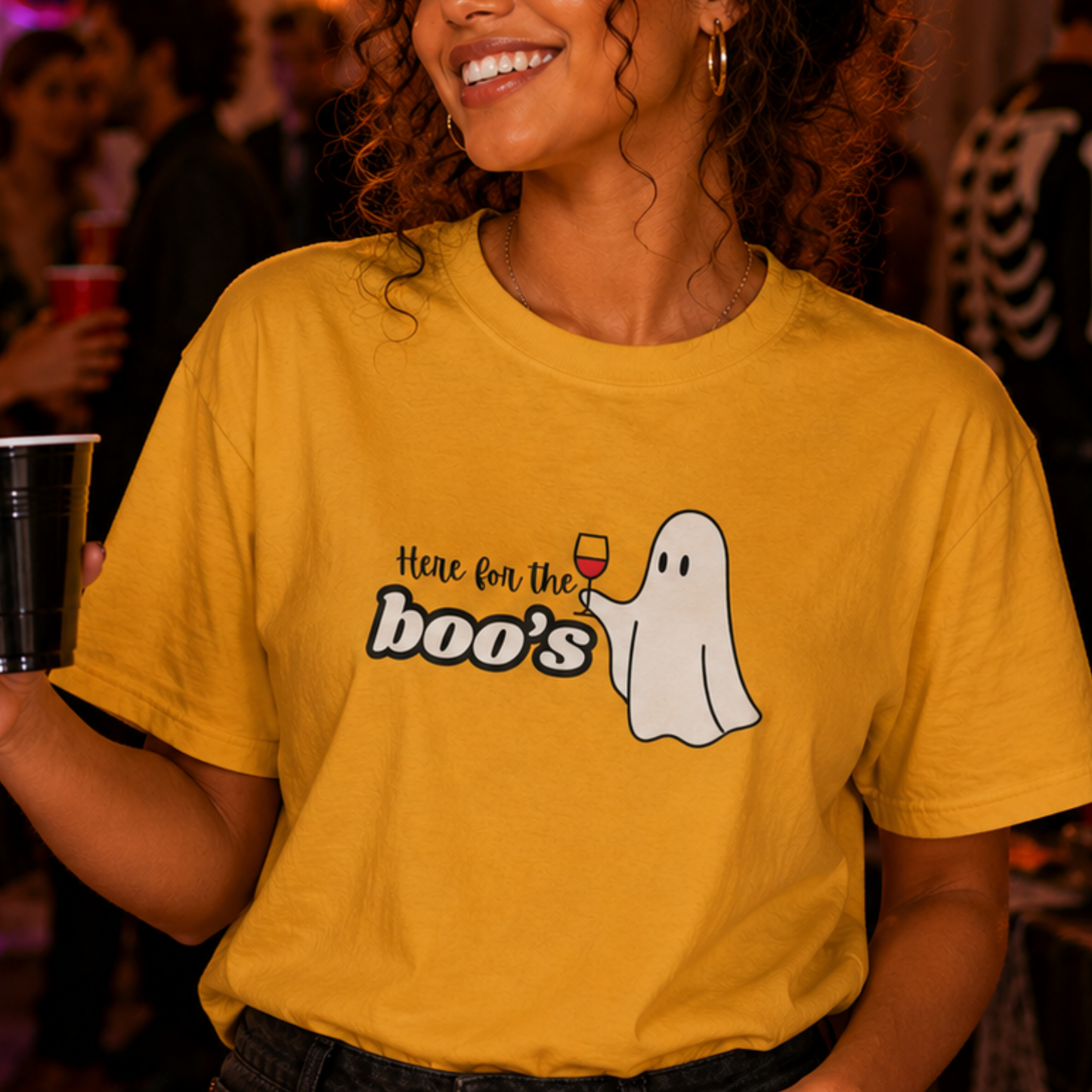 A woman at a party wearing a yellow graphic t-shirt with a ghost and the text 'Here for the boo's' holding a cocktail glass.