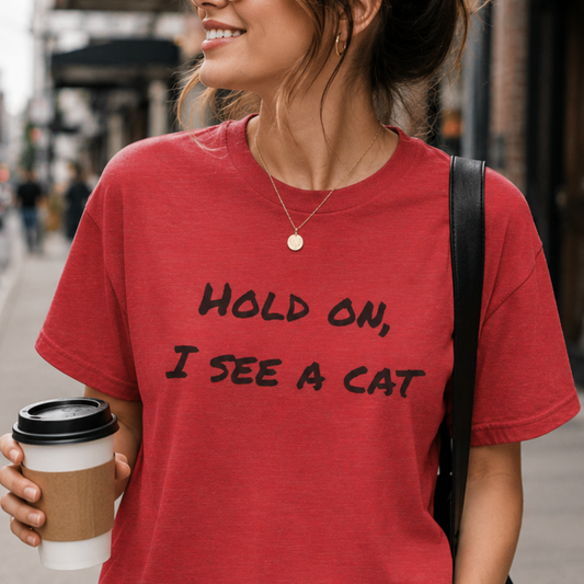 A woman wearing a red graphic t-shirt with the text 'HOLD ON, I SEE A CAT' in black handwritten-style lettering, holding a coffee cup on a city street.