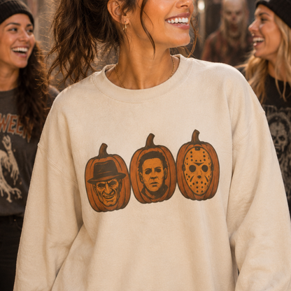 A woman wearing a cream-colored crewneck sweatshirt with a graphic of three horror movie pumpkin faces (Scary Face, Michael Myers, and Jason Voorhees) in orange and brown. The background shows a warm, out-of-focus indoor setting with other people smiling.