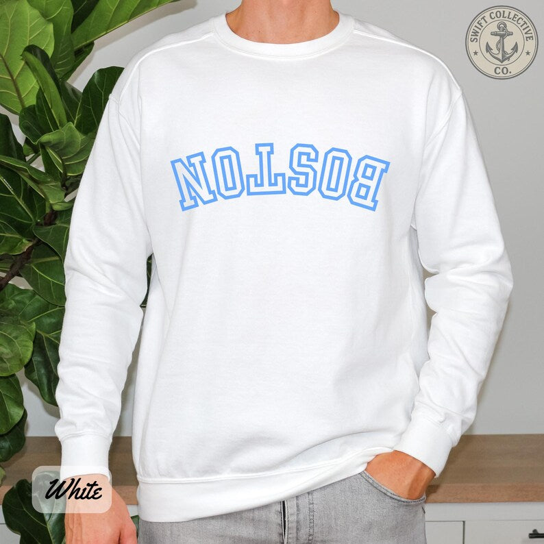A white crewneck sweatshirt with 'BOSTON' printed in blue arched varsity letters across the chest, worn by a model in front of a green plant.
