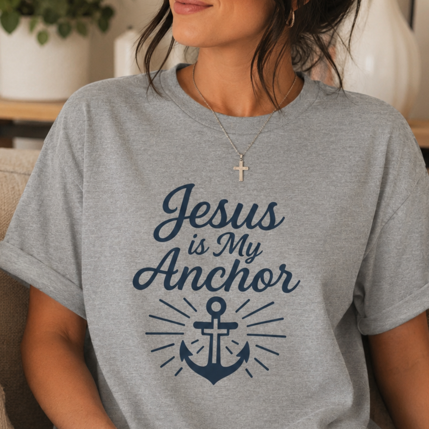 A woman wearing a heather gray t-shirt with a navy blue graphic that reads 'Jesus is My Anchor' above an anchor illustration, shown in a cozy indoor setting.
