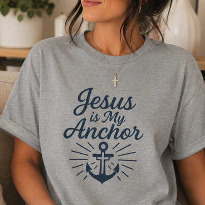 A woman wearing a heather gray t-shirt with a navy blue graphic that reads 'Jesus is My Anchor' above an anchor illustration, shown in a cozy indoor setting.