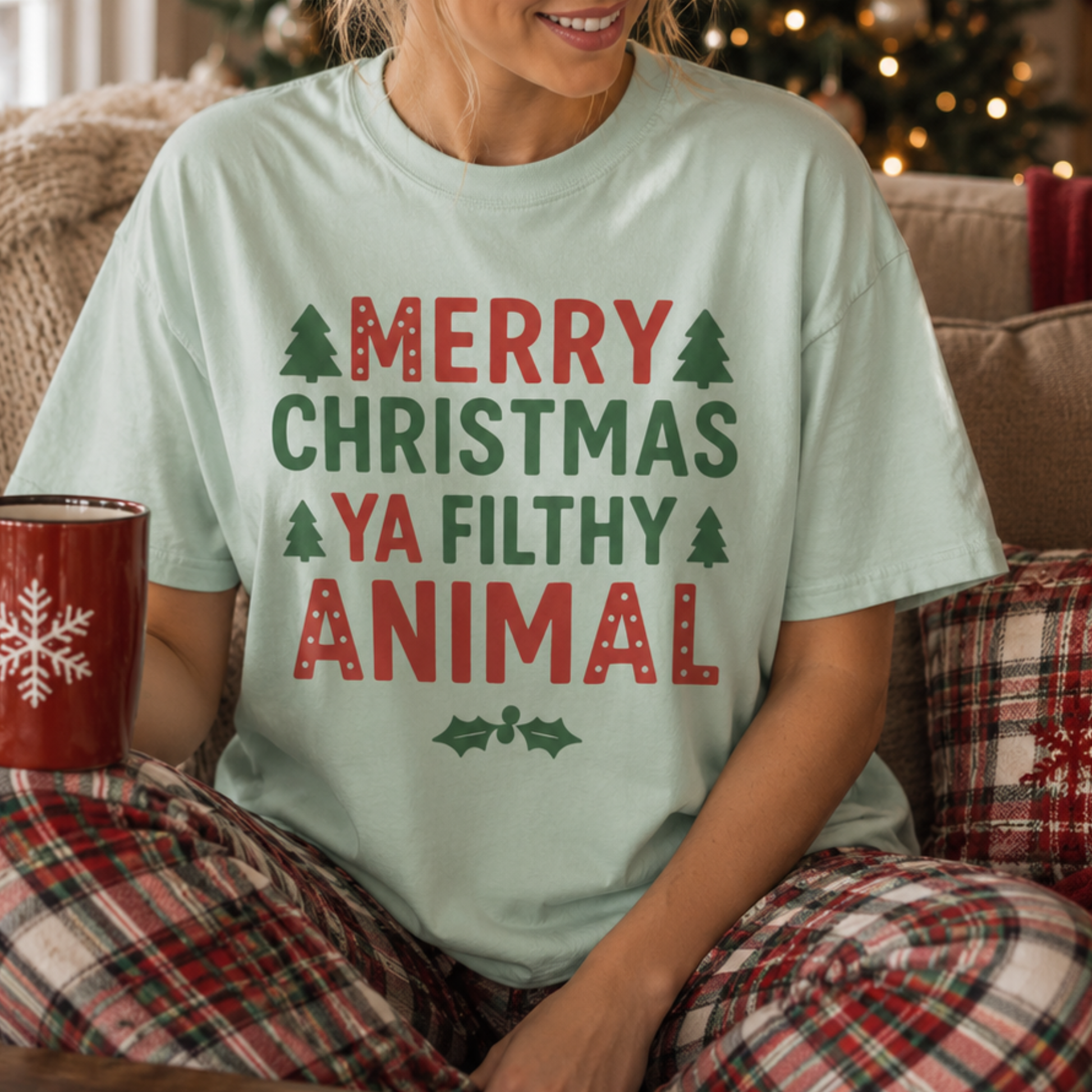 A woman wearing a light mint green graphic t-shirt with the text 'MERRY CHRISTMAS YA FILTHY ANIMAL' in red and green, paired with red and green plaid pajama pants, sitting in a cozy living room setting.
