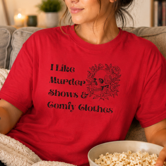 A woman wearing a red graphic t-shirt with black text that reads 'I Like Murder Shows & Comfy Clothes' next to a black skull and rose design. She is sitting on a couch holding a bowl of popcorn.