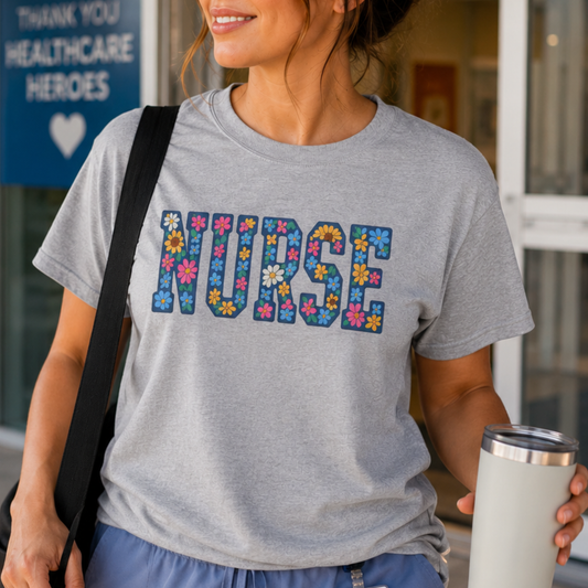 A woman wearing a heather gray t-shirt with the word 'NURSE' filled with colorful flowers, holding a travel tumbler and standing in front of a healthcare entrance.
