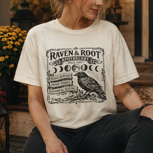 A woman wearing a cream-colored graphic t-shirt with a black 'Raven & Root Apothecary' design featuring a raven and moon phases, set in a warm, rustic outdoor environment.