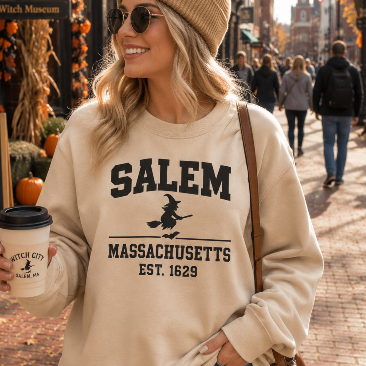 A woman wearing a beige crewneck sweatshirt with a black graphic that reads 'SALEM MASSACHUSETTS EST. 1629' and a witch riding a broomstick, standing on a cobblestone street in Salem.
