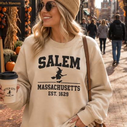 A woman wearing a beige crewneck sweatshirt with a black graphic that reads 'SALEM MASSACHUSETTS EST. 1629' and a witch riding a broomstick, standing on a cobblestone street in Salem.
