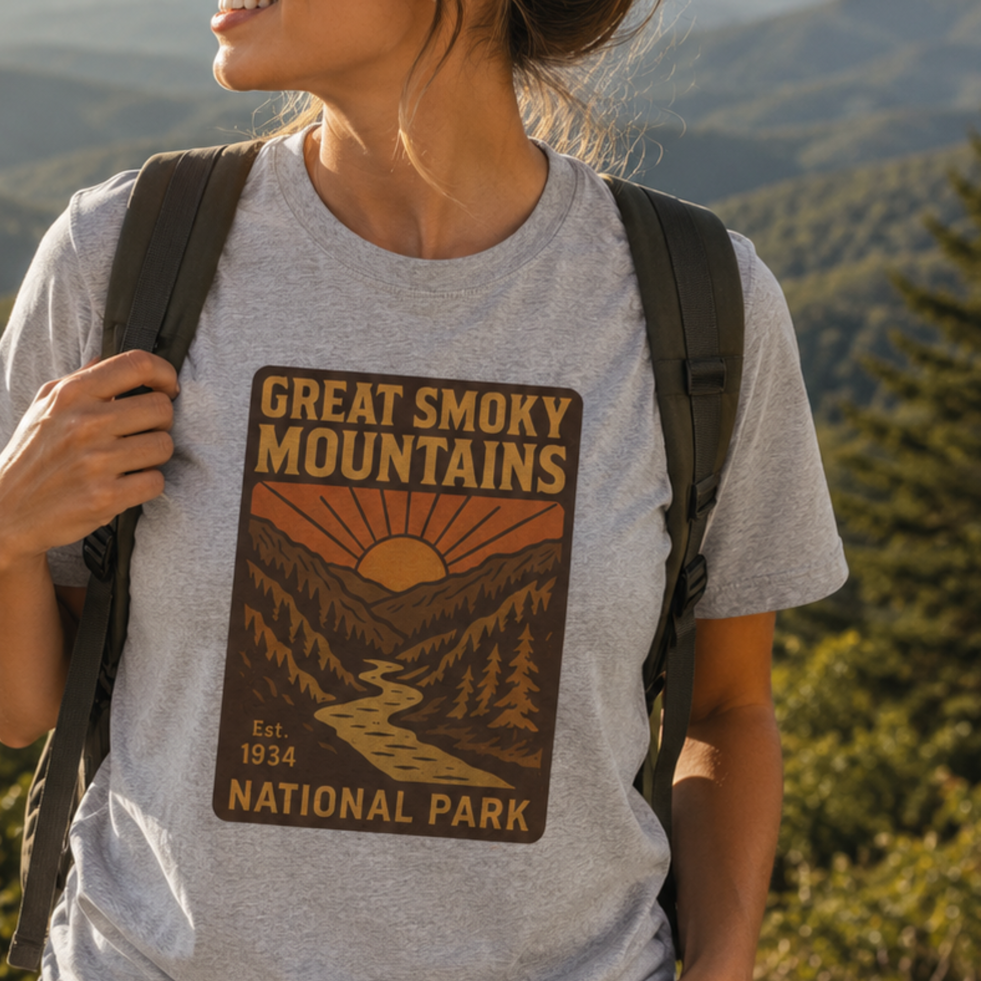 A woman wearing a heather gray t-shirt with a 'Great Smoky Mountains National Park' graphic print, carrying a black backpack outdoors with a blurred mountain forest background.