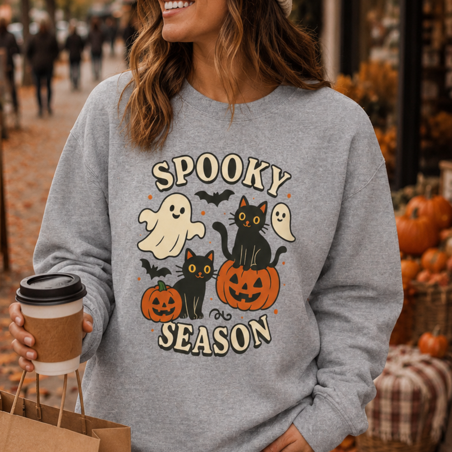 A woman wearing a heather gray crewneck sweatshirt with a 'Spooky Season' Halloween graphic, holding a coffee cup on an outdoor street.