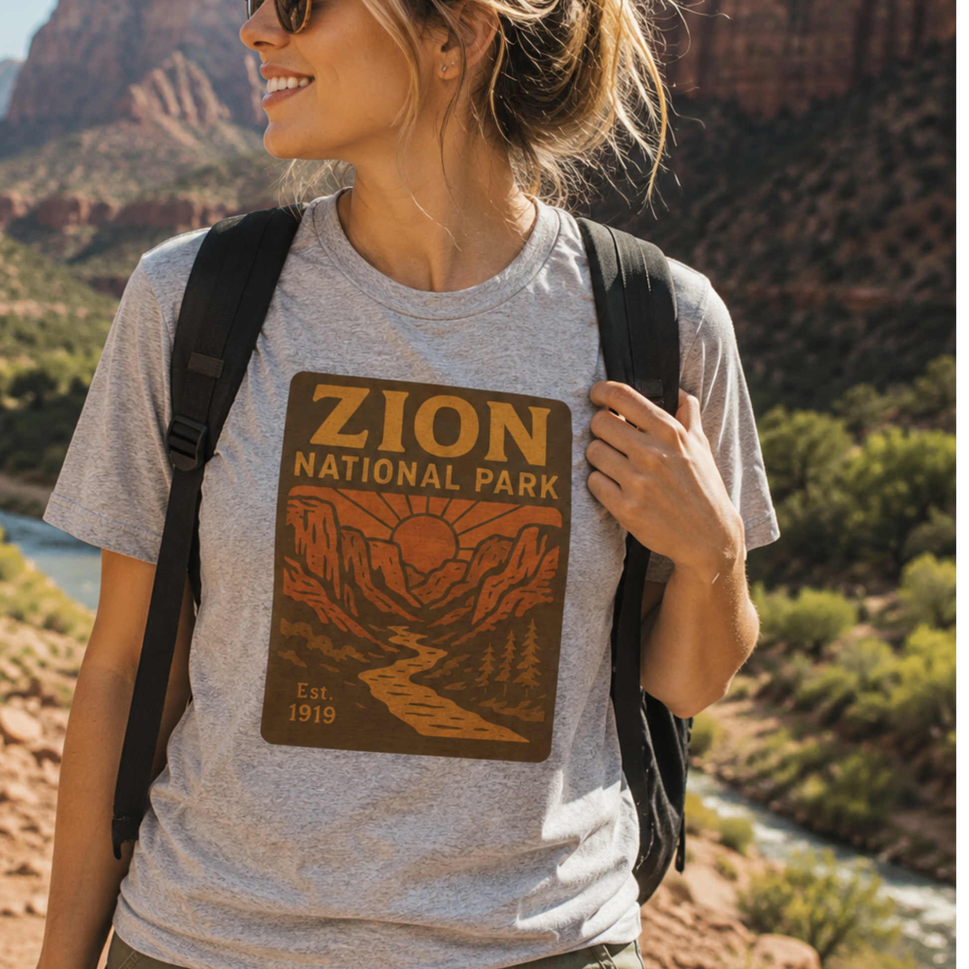 A woman wearing a heather gray t-shirt with a brown and orange 'Zion National Park' graphic print, carrying a black backpack in a scenic outdoor setting with red rock cliffs and a river.